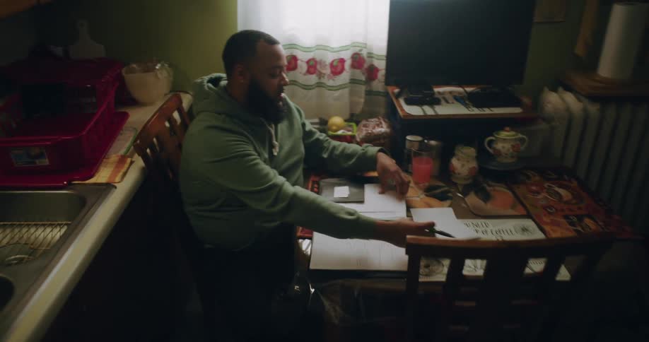 A man is sitting at a dining table in a cozy kitchen working on paperwork with a pen