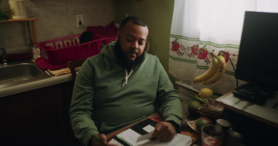 Man sitting in the kitchen at a table with a computer on it holds a business card and writes in a notebook
