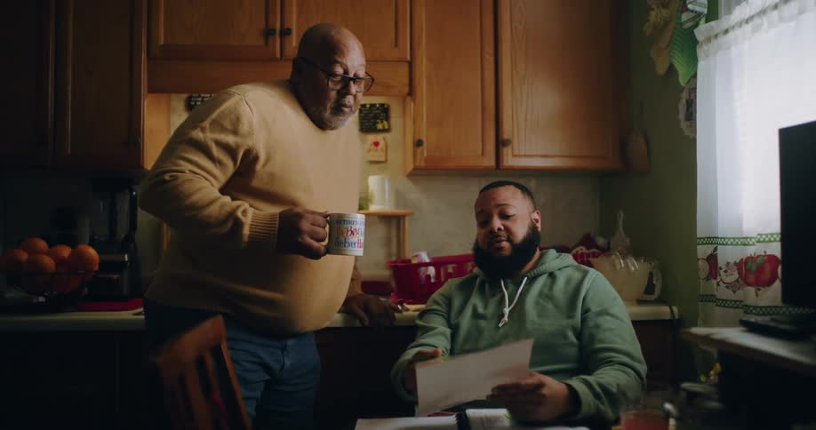 Two adult men in the kitchen with one of them showing a paper to the other