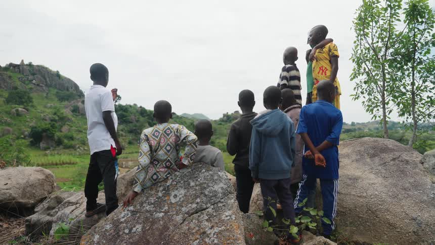 A group of children stands on rocks looking out over a lush hilly rural landscape with scattered trees and distant mountains