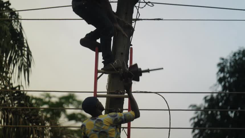 Two individuals are working on an electrical pole with one person climbing a red ladder and passing a component to other already positioned higher up on the pole