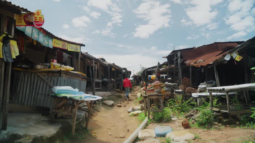 A person in a red hoodie walks down a narrow dirt path flanked by makeshift market stalls with corrugated metal roofs and various items for sale under a partly cloudy sky