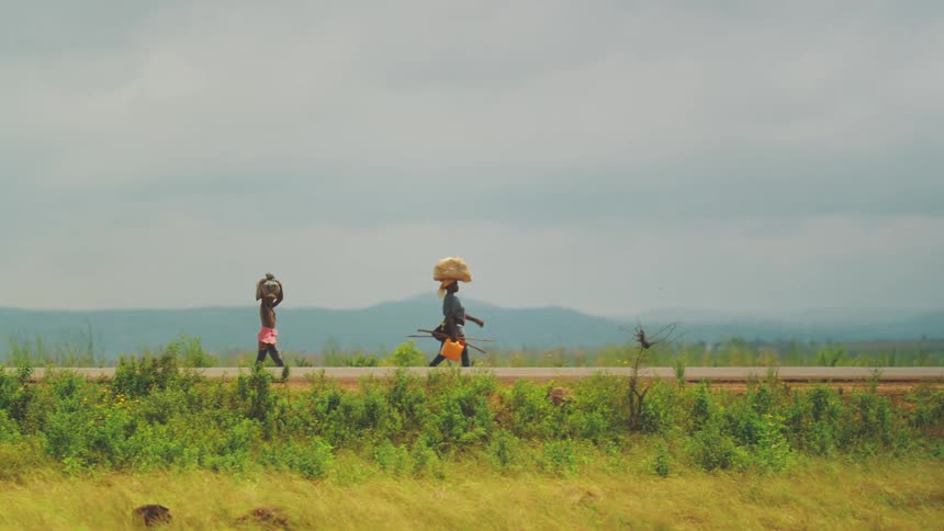 Two people walking on an outdoor street surrounded by vegetation and carrying bags suspended on their heads