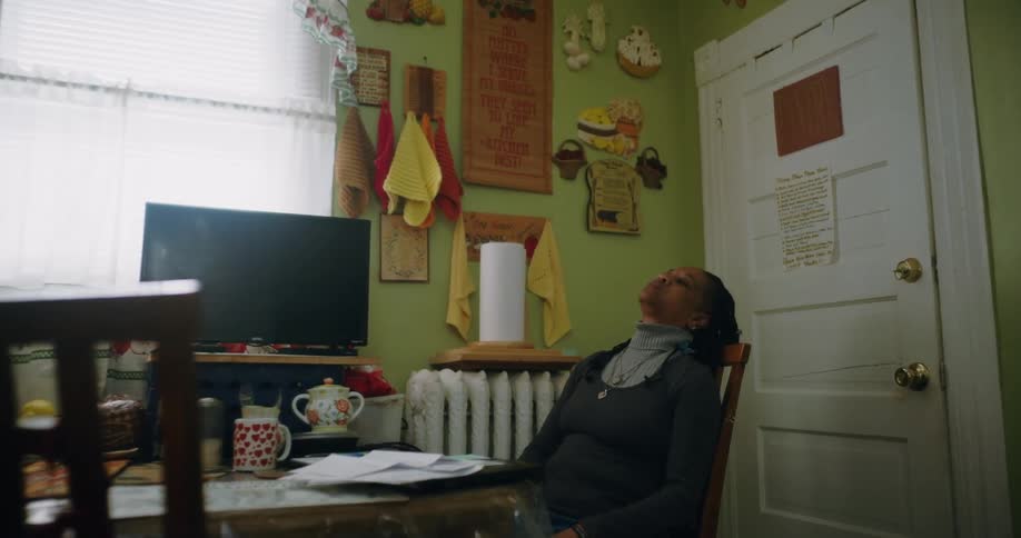 A woman sits at a table in a cluttered kitchen looking upwards with her hands clasped in her lap