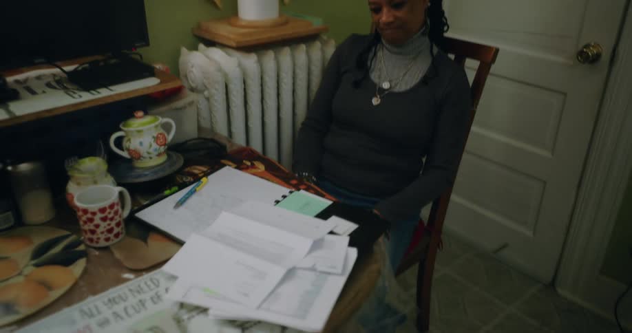A woman is sitting at a dining table in a cozy kitchen working on paperwork with a pen