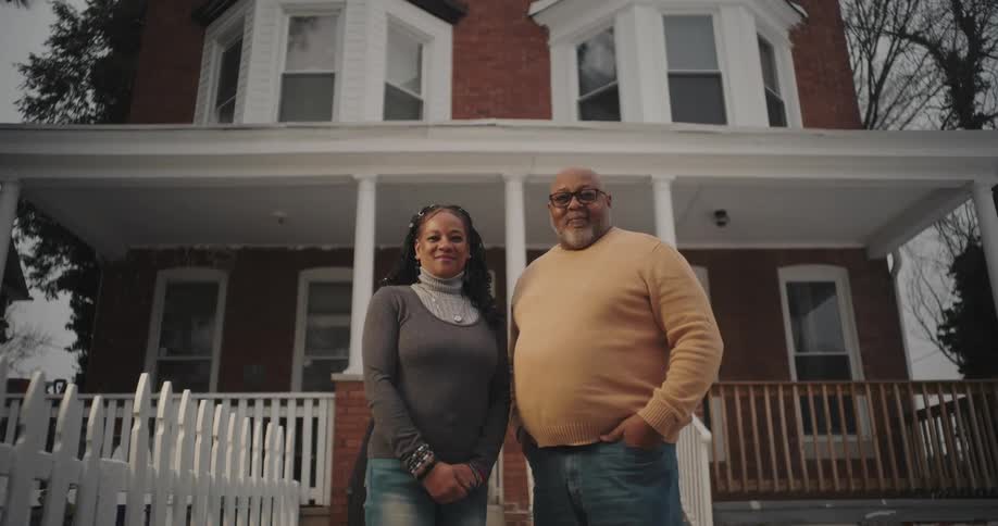 Man and woman with happy facial expressions are posing in front of a house