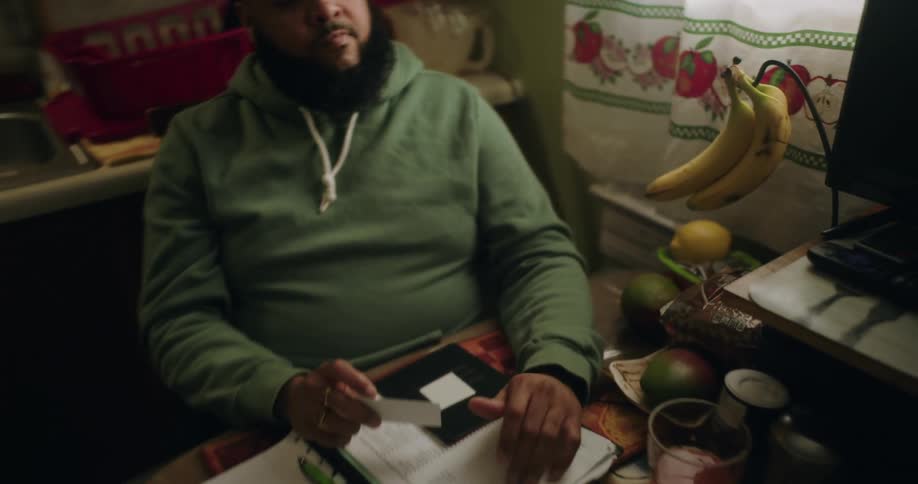 A person in a green hoodie sits at a kitchen table with a notebook pen and various papers appearing to work on taxes