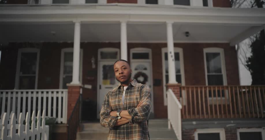 A man standing in front of a twostory brick house with a white picket fence and a wooden porch