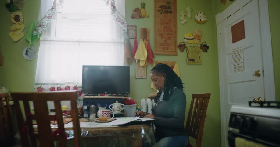 A woman is sitting at a dining table in a cozy kitchen working on paperwork with a pen