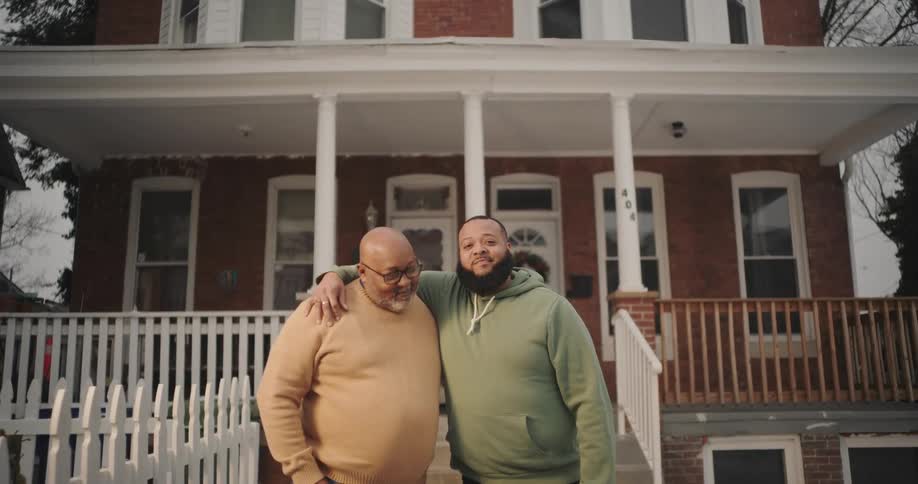 Father and son are standing in front of a twostory brick house with a white picket fence and a wooden porch