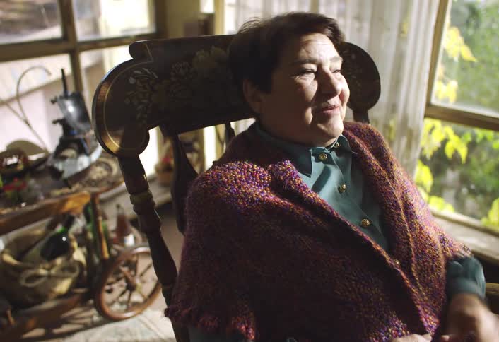 A smiling woman with short dark hair wearing a shawl sits in an ornate rocking chair and appears to be talking to someone off screen