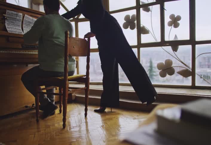 A pianist playing an upright piano with sheet music in front of them while woman dressed in black stands beside him gesturing expressively giving instructions with a large window featuring decorative floral metalwork and a cityscape in the background