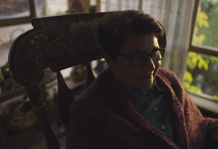 A woman sits in a wooden rocking chair with floral carvings wearing a maroon knitted shawl near a window with yellow foliage visible outside