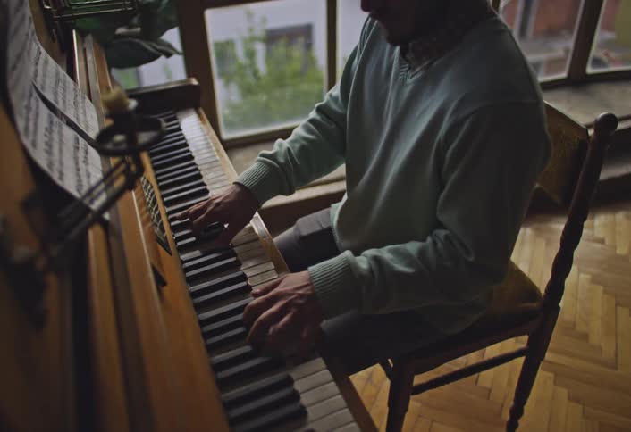 A person wearing a light green sweater is playing a wooden piano with sheet music placed on the stand in a room with large windows showing a view of greenery outside