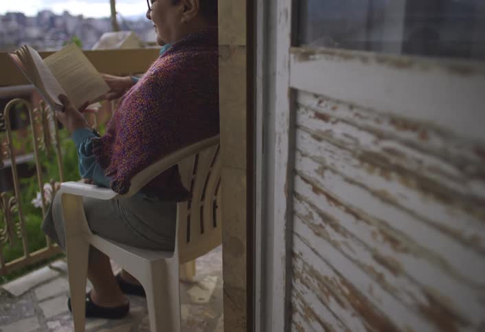 Elderly woman sitting on a plastic chair on the porch reading a book