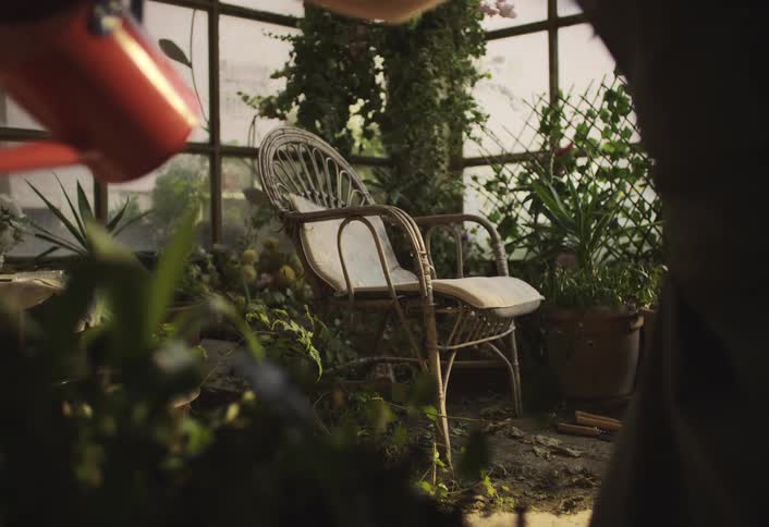 A person waters plants in a sunlit greenhouse with a wicker chair cushion and various potted plants and vines