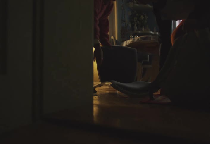 A teenage boy knocks over a chair while grabbing a backpack in a dim dining room picks it up with a womans help and then runs away