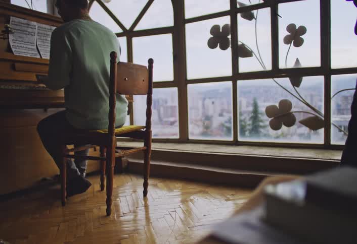 A person is seated at a piano playing music while another person stands nearby in a room with large windows and wooden flooring