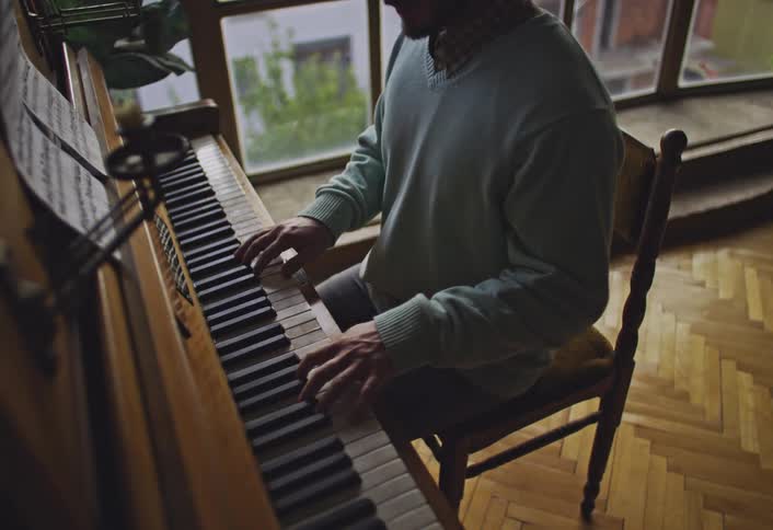 A man sits down and begins to play an antique piano in a room with parquet flooring and a window in the background
