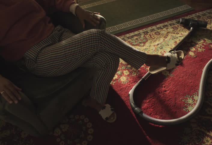 A woman is sitting on a chair with legs crossed wearing checkered pants and white slippers next to a vacuum cleaner on a red patterned carpet