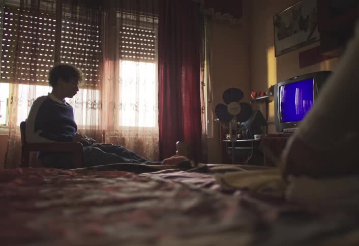 A teenager is playing video games in a dimly lit room while someone is folding laundry in the bed