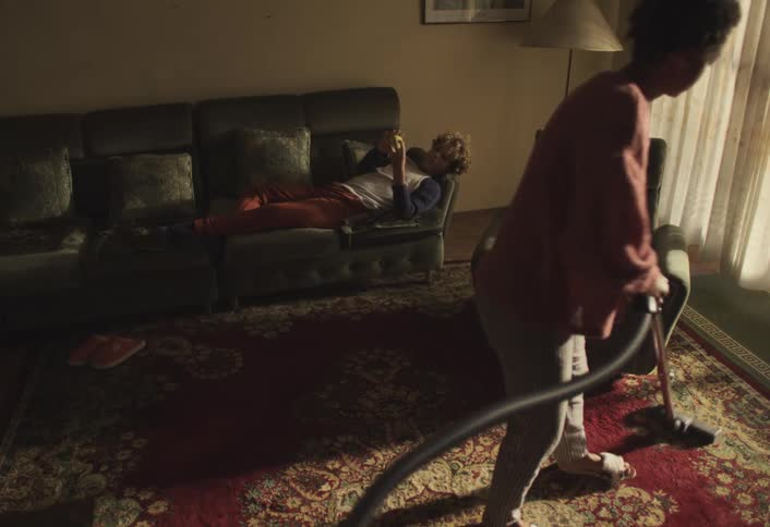 Youth lying on the couch plays with a ball while a woman vacuums the living room