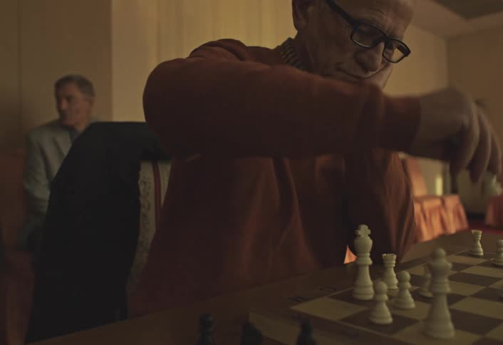 An elderly man engaged in a game of chess in a dimly lit room with a row of empty chairs and sheer curtains in the background