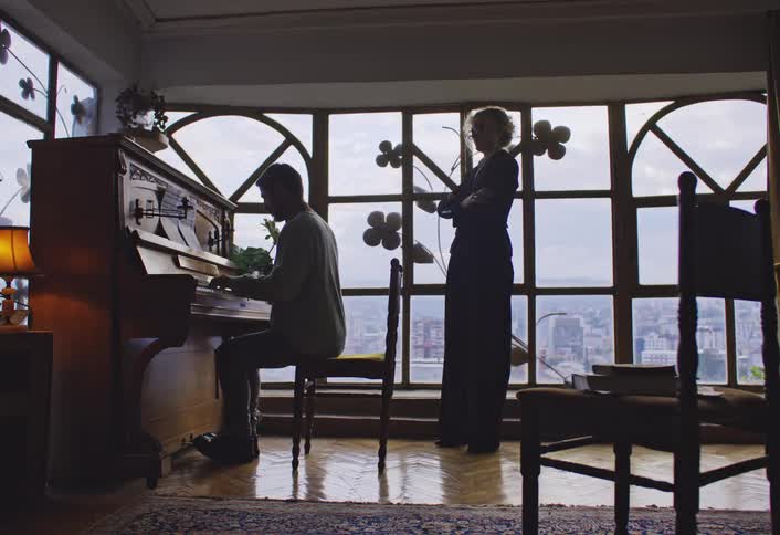 A person is playing a piano in a room with large windows decorated with flowershaped decorations while another person sits nearby
