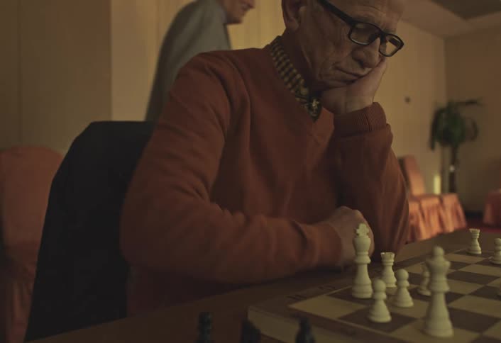 An elderly man engaged in a game of chess in a dimly lit room with a row of empty chairs and sheer curtains in the background