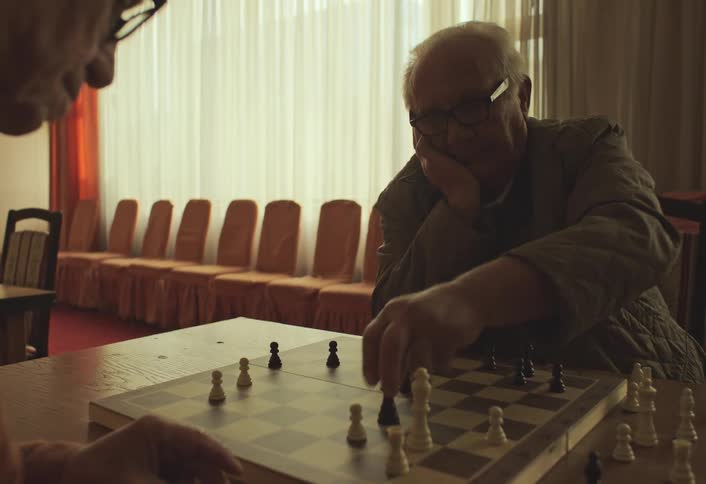 Two elderly individuals are engaged in a game of chess in a dimly lit room with a row of empty chairs and sheer curtains in the background