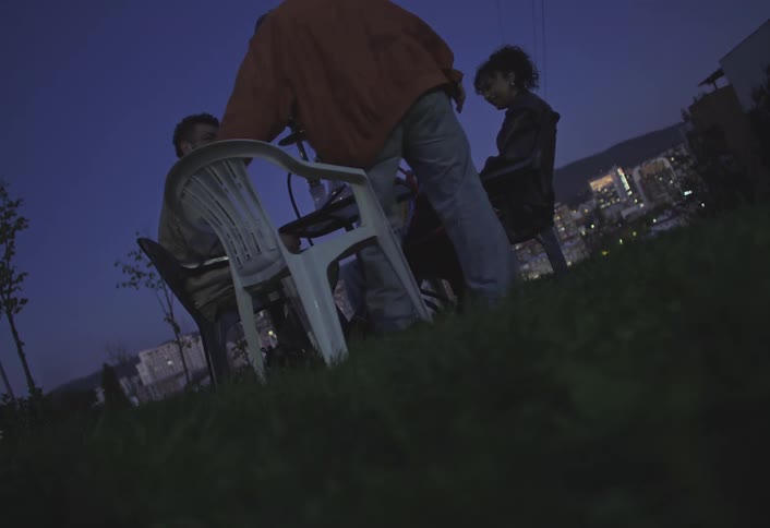 Four friends are seated on plastic chairs in an outdoor setting during twilight with a cityscape visible in the background