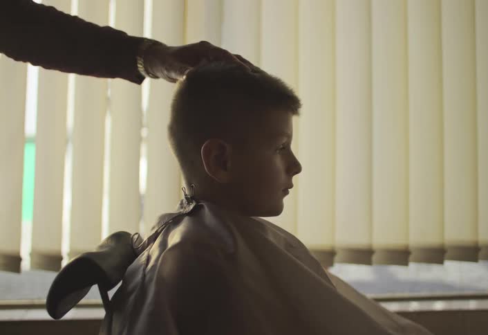 A child sits in a barber chair getting a haircut from a stylist in a salon with venetian blinds