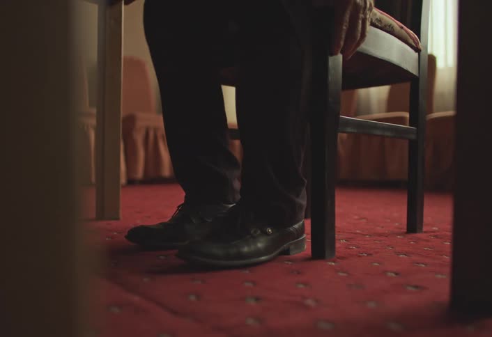 A person wearing black shoes and dark pants sits on a chair in a room with red carpet flooring
