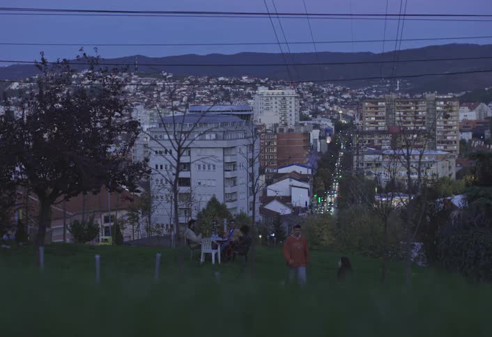 Group of people sitting outdoors on a hill overlooking the city at dusk and a couple walking away
