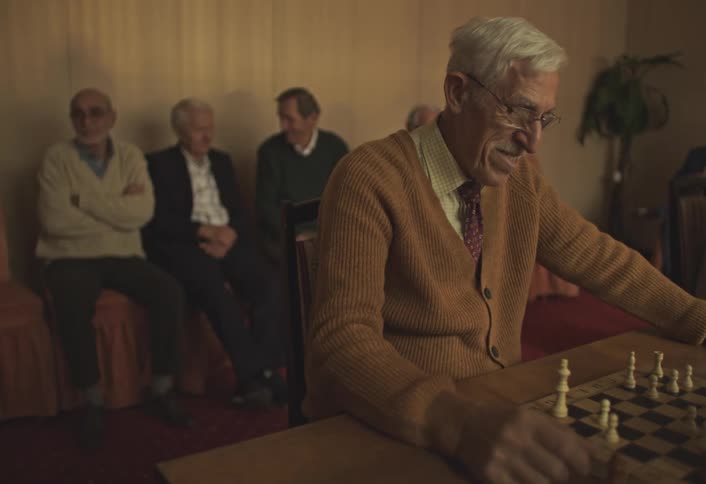 Elderly man sitting playing chess and other men sitting in armchairs just behind him in the same room