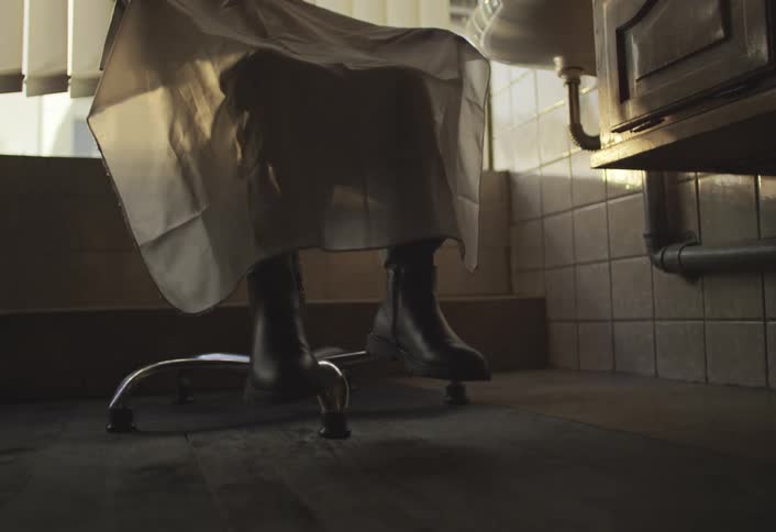 Person sitting on a swivel stool wearing dark boots and covered by a hairdressers cape