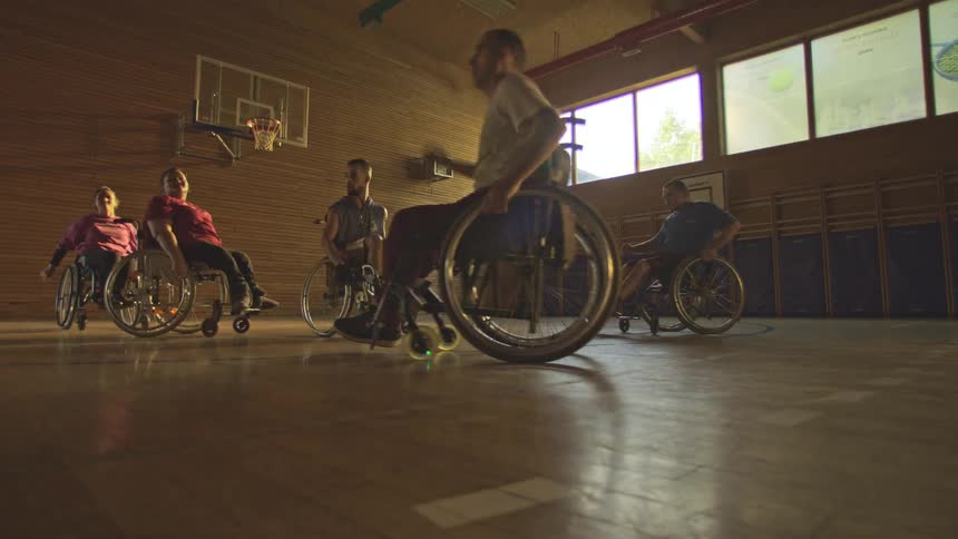 Players in wheelchairs play basketball in a gym with wooden walls and natural lighting