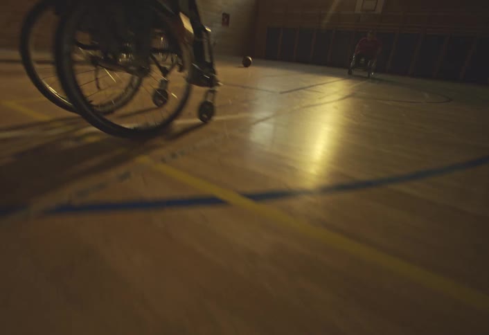 People in wheelchairs play basketball in an indoor gymnasium with sunlight streaming through large windows