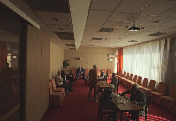 A group of elderly people gathered in a room with chairs arranged around them and some playing chess