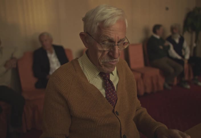 Elderly man in brown sweater and glasses looks down with concentrated expression while other people sit in the background