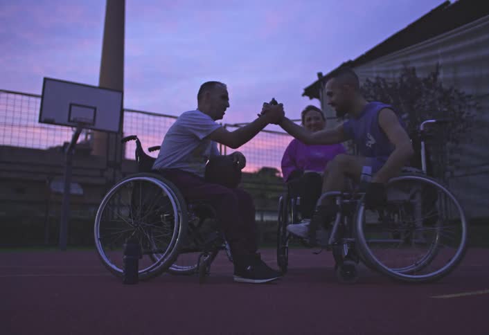 Three individuals in wheelchairs are conversing on an outdoor basketball court at dusk