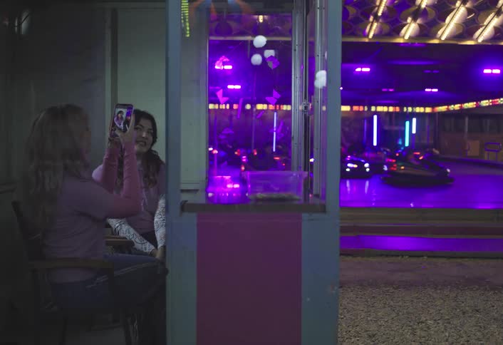 Two women sit taking a photo near a booth at an amusement park with illuminated bumper cars in the background