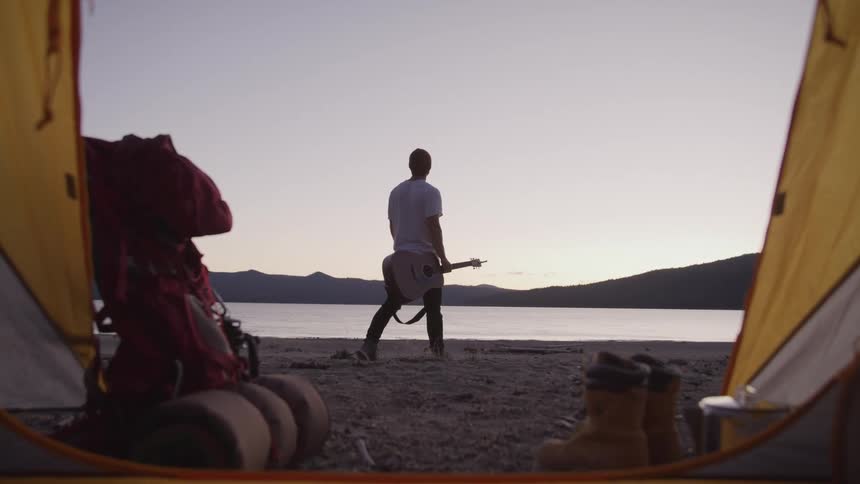 View from inside a tent of a man in a beret holding a guitar as he walks along the beach at dusk