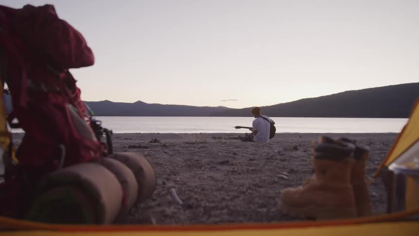 View from inside a tent of a man playing guitar on the beach at dusk with backpack and boots in the foreground