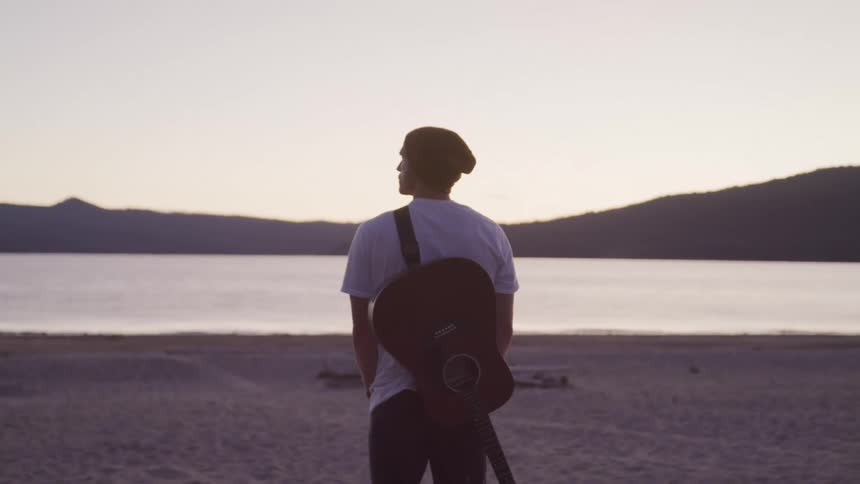 Man in a beret carries a guitar on his back as he watches the sea at sunset