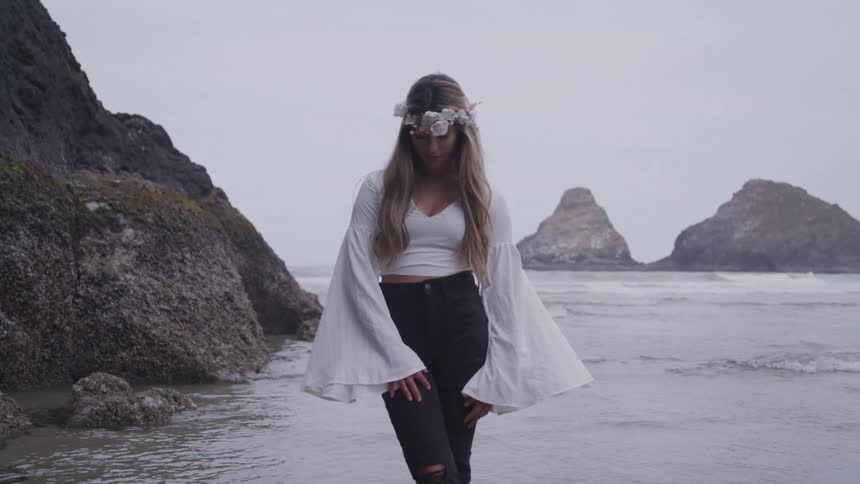 A woman with long hair wearing a white longsleeved top and black pants stands on a rocky beach with the ocean and two large rock formations in the background