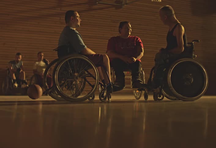 Group of players in wheelchairs chat on a basketball court with a ball on the floor