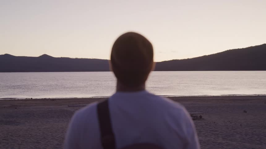 Man in beret looks out to sea at sunset with mountains in the background