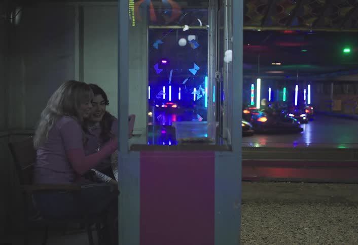 Two women sit in a booth taking selfies while a brightly lit bumper car arena with several bumper cars in motion is visible in the background