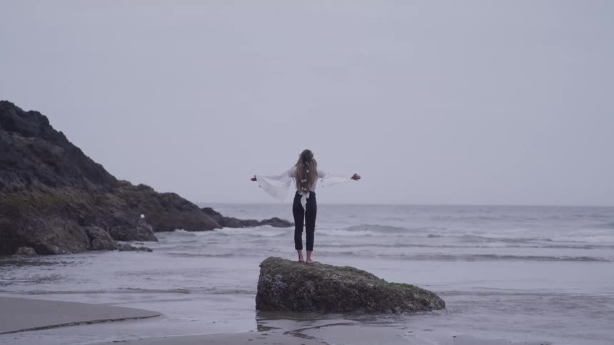 Barefoot woman spreading her arms on top of a rock on the beach with the sea in the background on a cloudy day